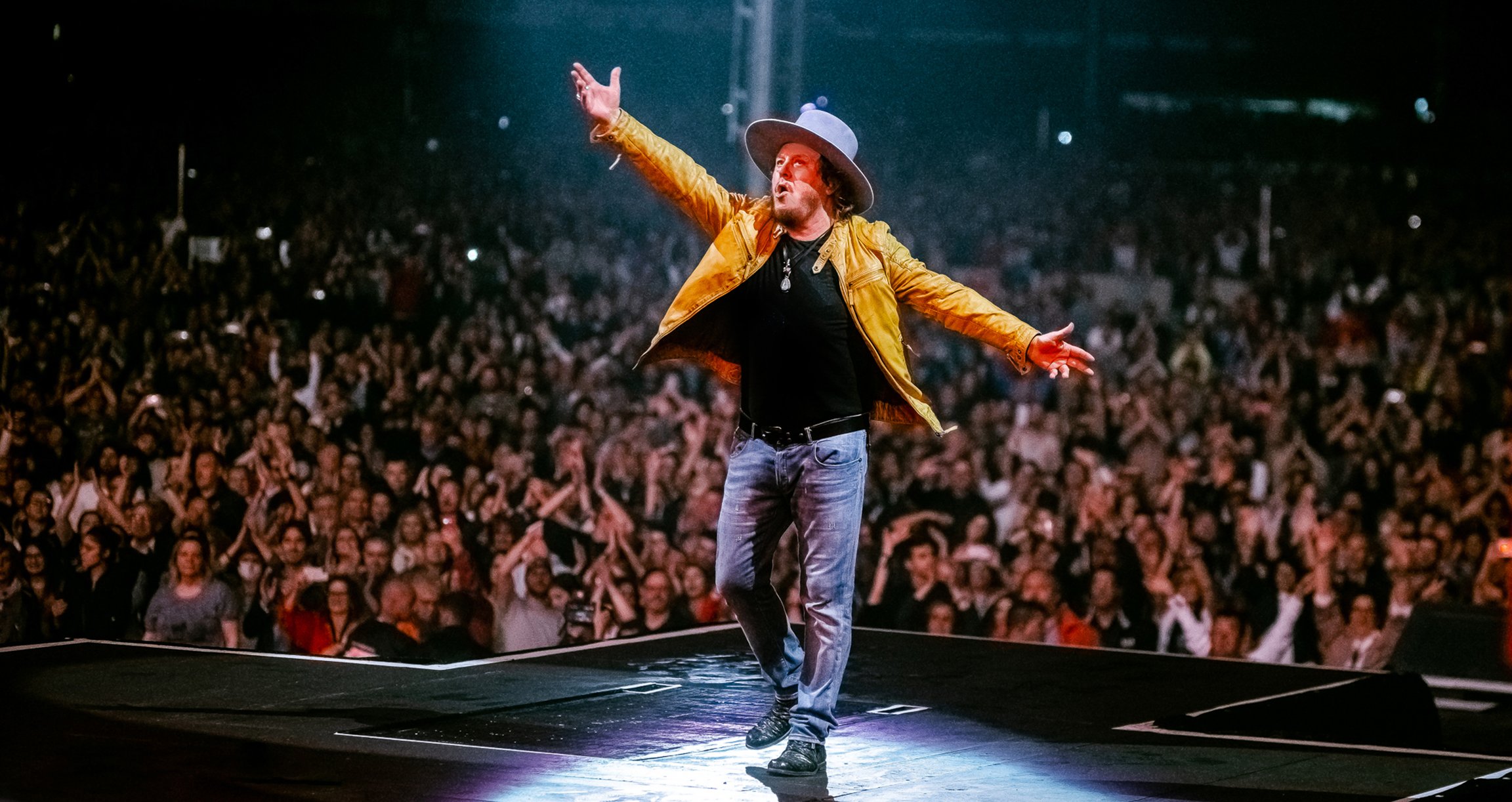 Zucchero in gelber Jacke auf der Bühne bei einem Live-Konzert, im Hintergund jubelt das Publikum.