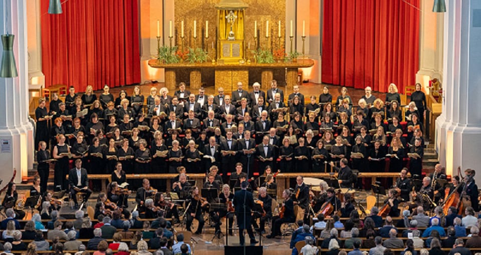 Ein Chor und ein Orchester präsentieren sich in einer Kirche, die von roten Vorhängen umrahmt wird, und erzeugen eine feierliche Stimmung.