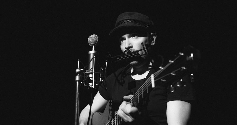 Ein schwarz weiß Bild auf dem ein Mann mit einem Hut sitzt eine akustische Gitarre spielt