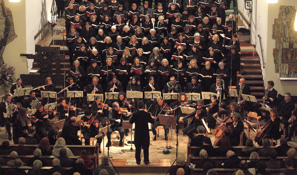 Ein Chor und ein Orchester führen in einer Kirche ein musikalisches Stück auf, vor Ihnen steht ein Dirigent.