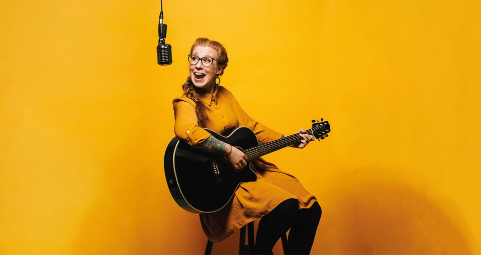 Eine Frau in einem gelben Kleid hält eine Gitarre in der Hand und singt dabei in ein Mikrofon 