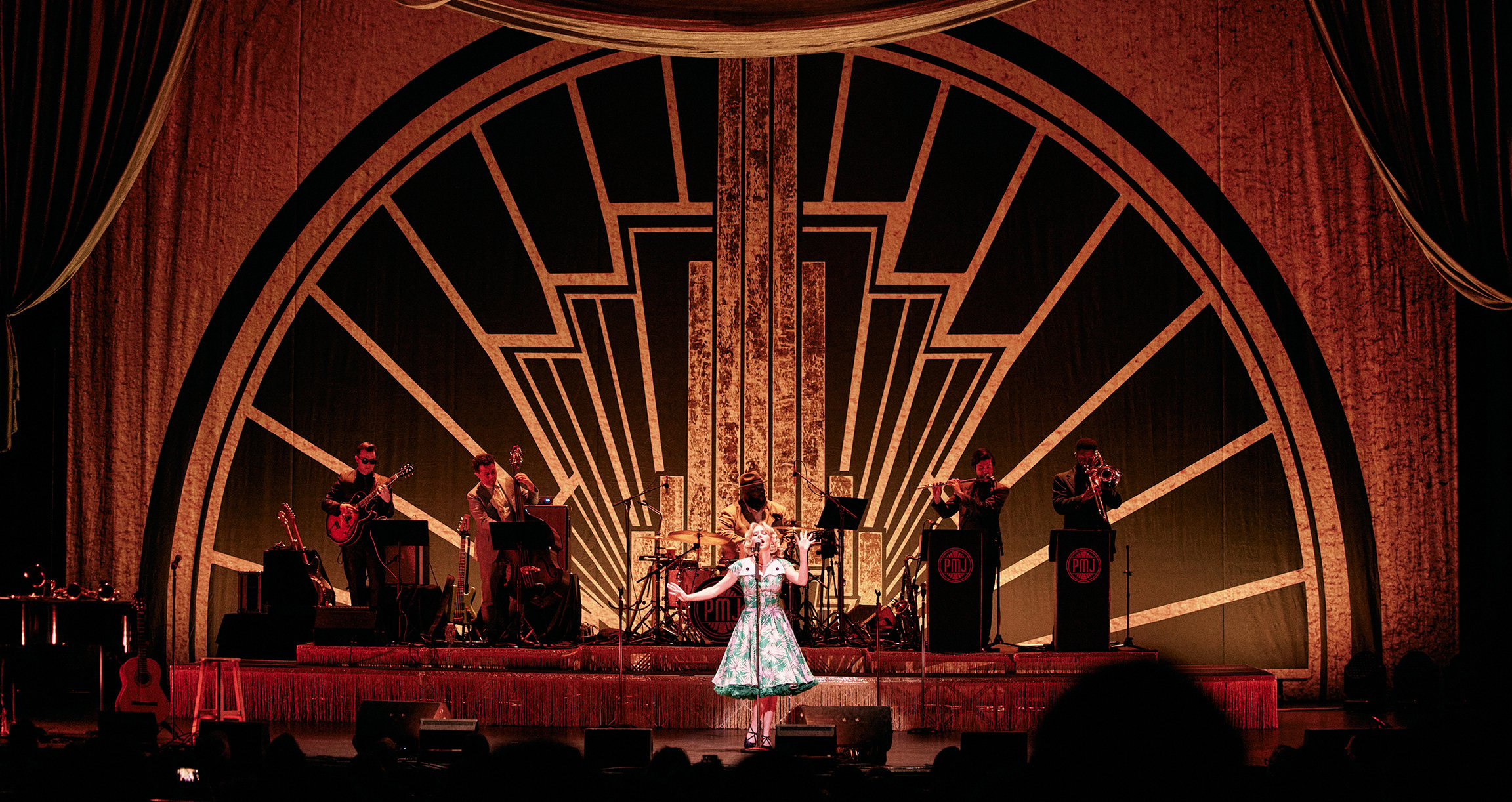 Auf der Bühne steht eine Frau in einem eleganten Kleid, umgeben von einer Band, die für das Publikum spielt.