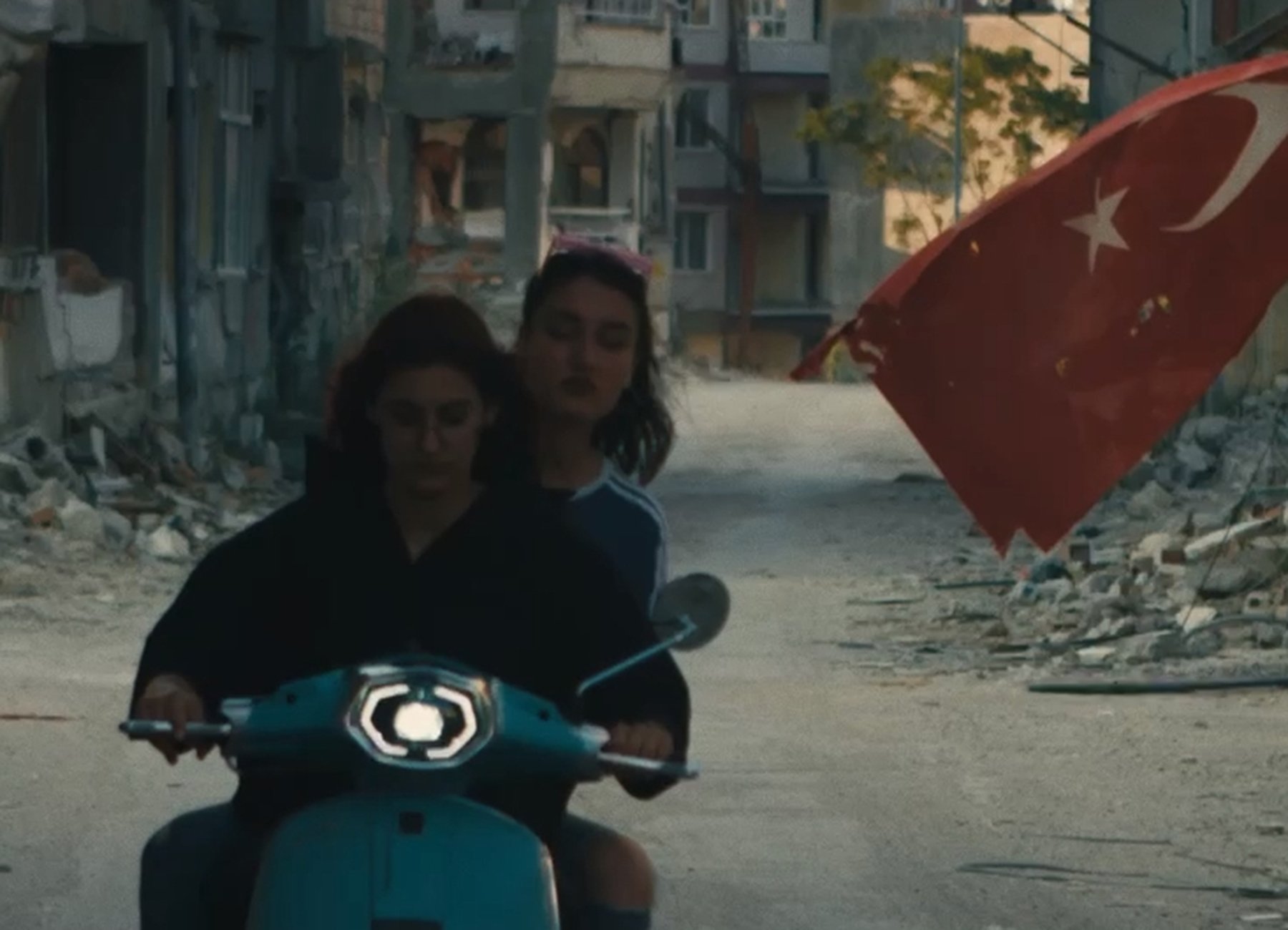 Zwei Personen fahren mit einer türkischen Flagge im Hintergrund auf einem Roller durch eine Gasse 