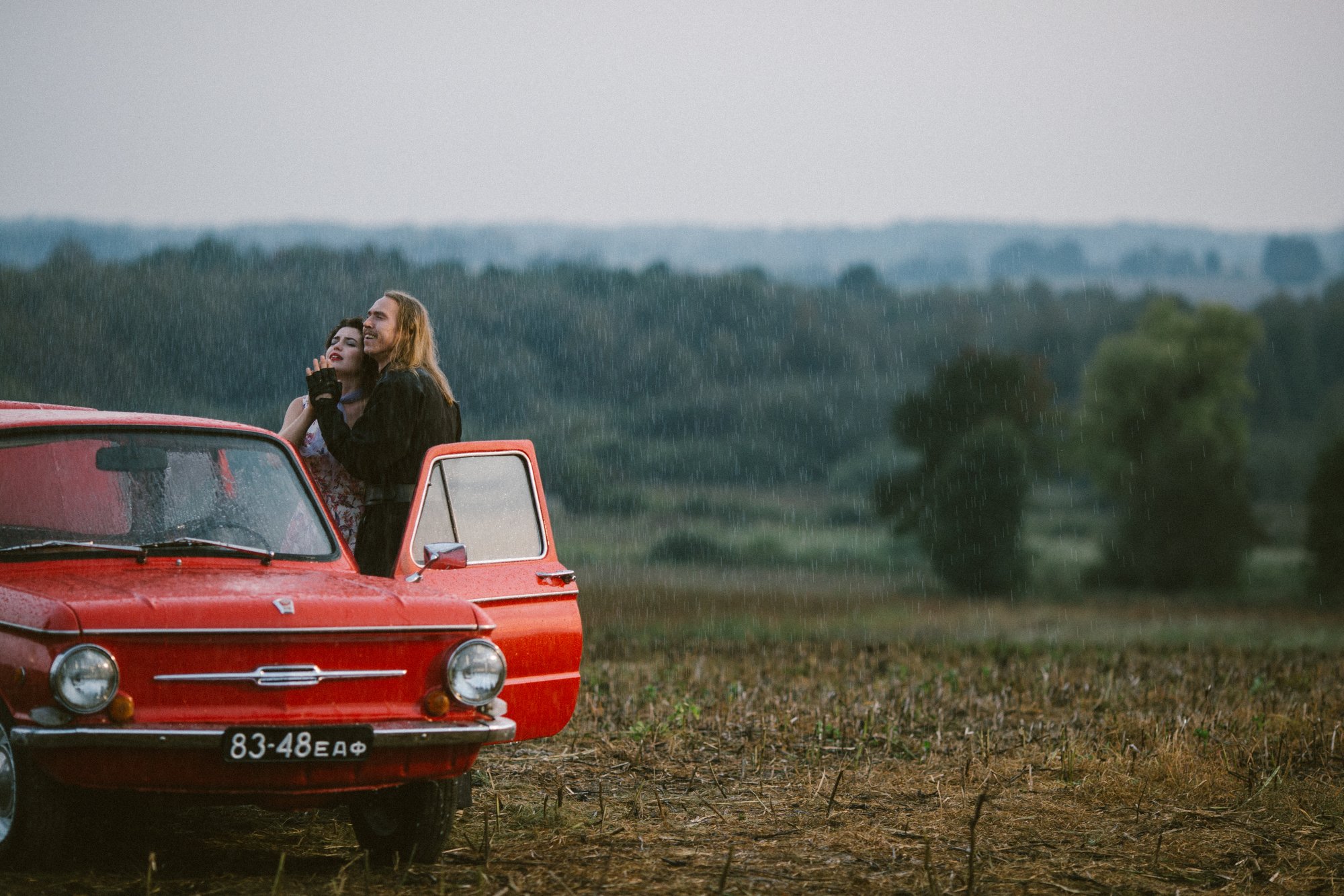 Ein Mann und eine Frau stehen Arm in Arm im Regen, vor einem Auto 