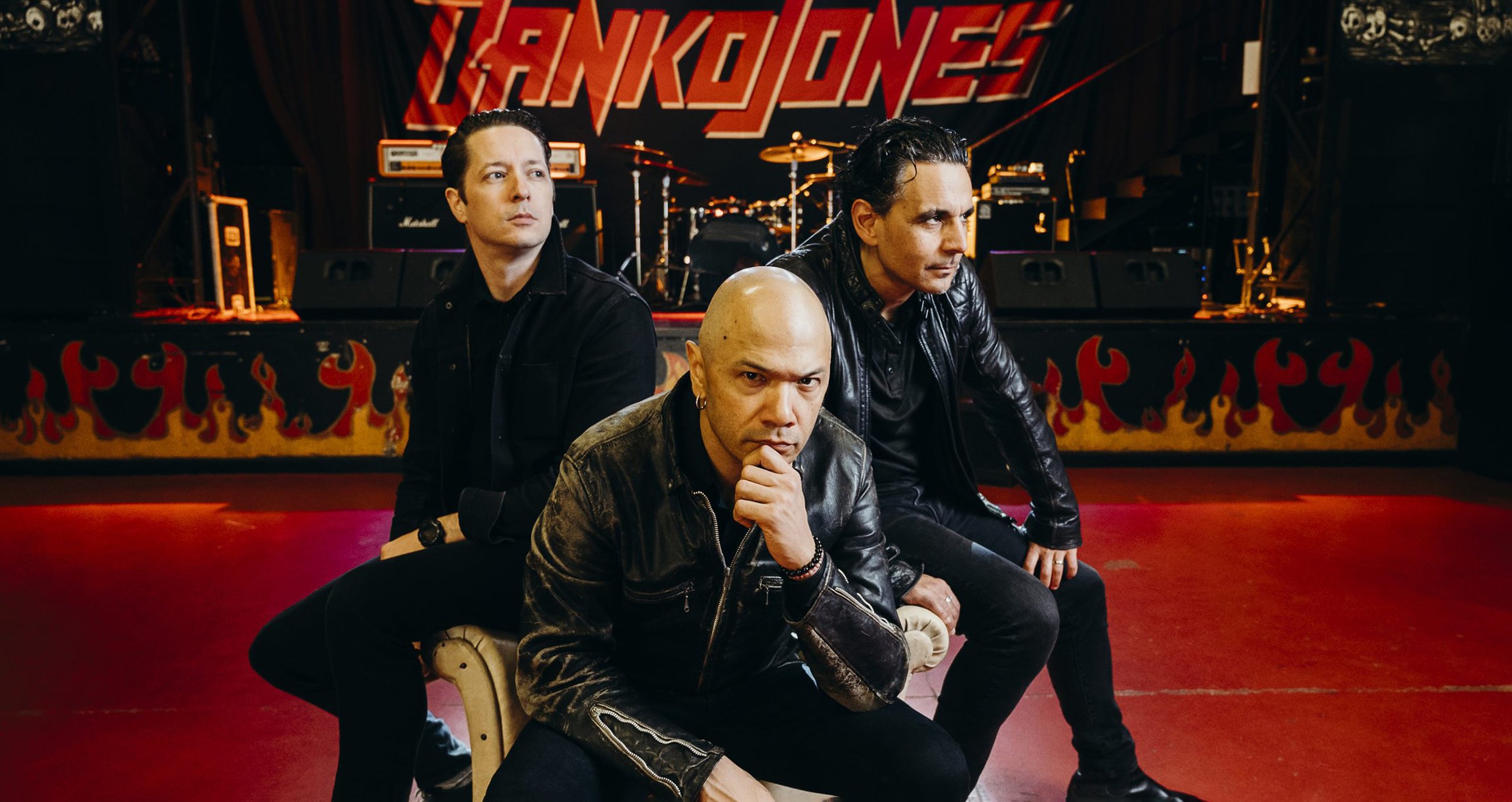 Danko Jones sitzen in dunklen Outfits in einer Konzerthalle vor einer dunklen Bühne