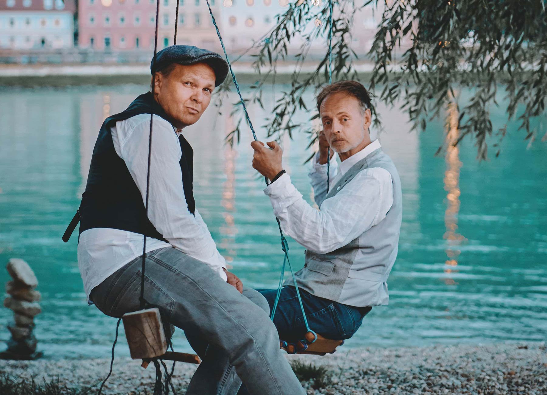 BlöZinger sitzen vor einem Fluss auf Schaukeln und gucken in formeller Kleidung in die Kamera